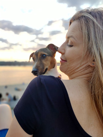 A young blonde woman holds a Jack Russell dog on her shoulder. The sky with clouds is on the back blurred backgroundの写真素材