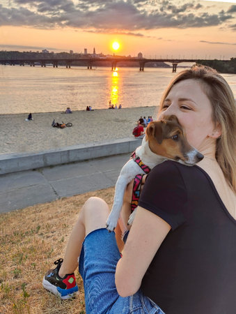 A blonde girl holds a dog of the Jack Russell terrier breed in her arms and sits on the bank of the river at sunset. Rear viewの写真素材
