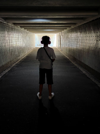 Outline of a teenage boy wearing headphones standing in an underpass facing a bright exit. Rear view in perspectiveの写真素材