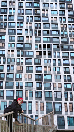 A young Caucasian teenager in a cap and jacket stands against a skyscraper. The building has many...