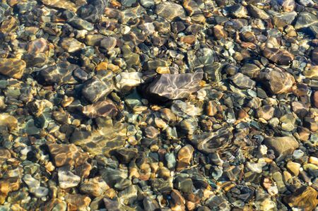 River pebbles under a thin layer of transparent water, illuminated by the sun.