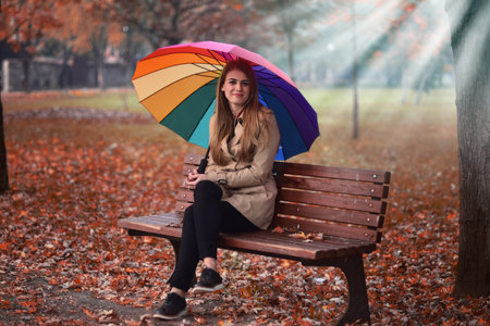 Young beautiful girl sitting on the bench in the autumn park with a umbrella.の写真素材