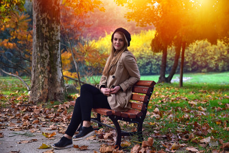 Young girl sitting on bench in public park at autumn. Girl wearing cream color coat and black beret relax in nature.の写真素材