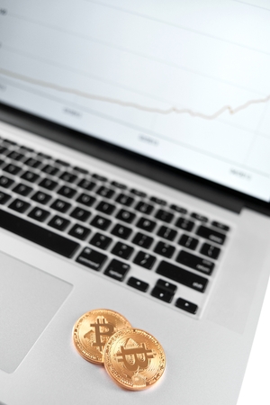 Two golden bitcoins placed on silver laptop with financial chart on its screen.の写真素材