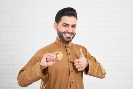 Smiling handsome muslim model in traditional Islamic clothing posing with golden bitcoinの写真素材