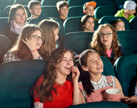 Happy girls laughing by watching interesting movie in the cinema. They smiling, looking satisfied. There are many other emotional children on the background.の写真素材