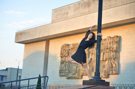 Ivano-Frankivsk, Ukraine - 1 June 2015 : Joung girl is dancing ballet on the street near lamp pillar. Intresting buildings facade on background. Model wearing balck dress and having long curly hair.のeditorial素材