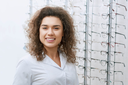 Portrait of young smiling curly ophthalmologist posing near stand with eyeglasses. looking frienly...