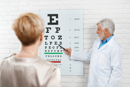 Photo of old ophthalmologist is cheking patients vision. Doctor working in medical laboratory near table for vision cheking. Wearing white medical uniform. Looking expirienced and skilled.の写真素材
