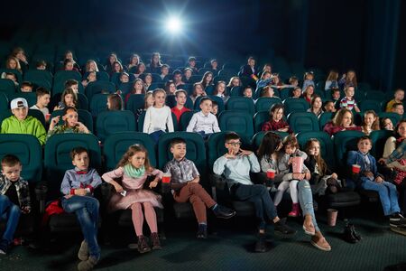 Frontview of people sitting in the cinema hall and watching movie. Boys and girls watching interesting movie and looking very emotional, frightened and exited. Children wear colorful trendy clothes.の写真素材