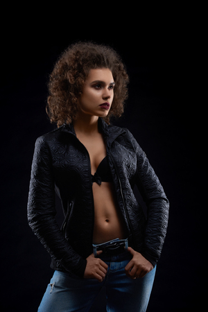 Sexy woman posing in black jacket and blue jeans. Looking beatiful,confident, charismatic. Having pretty face features, many little curles and light day make up. Studio black background.の写真素材