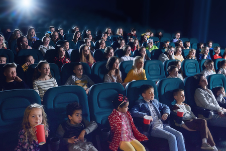 Crowd of people sitting in comfortable seats in modern movie theatre. Children, teens, adults watching  film and sipping fizzy drinks. Concept of cinematography and entertainment.の写真素材