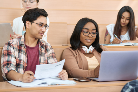 Groupmates working with a laptop in modern spacy university classroom with cascade wooden desks. Looking satisfied and concentrated. Coworking for educational scientific project.の写真素材