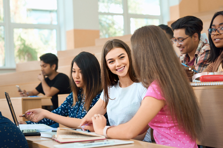 Smiling students talking during lesson. Sitting in modern university classroom with wooden desks on groupmates background. Guys reading, studuing, writing. Boys and girls wearing casual clothes.の写真素材
