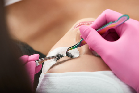Professional is making long lashes for a client wearing pink hygienic gloves. Master is using different types of tweezers. Woman lying on special coach with closed eyes during the process.の写真素材