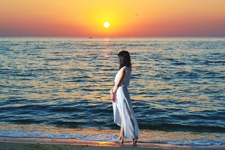 Girl wearing white summer dress standing on beatiful sunsets background. Looking on evenings deep blue sea and waves. Amazing, colorful sunset. Standing on sandy beach.の写真素材