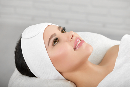 Smiling woman lying on white cosmetological coach wearing headbandage. Waiting for beaty salon procedures and professinal master. Smoth, young, healthy skin. Beatiful face.の写真素材