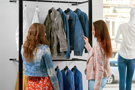 Backview of two girls buying clothes in the shop. Choosing fashionable jackets and colorful saturated shirts. Standing on white wall background. Interested clients ready to make choice.の写真素材