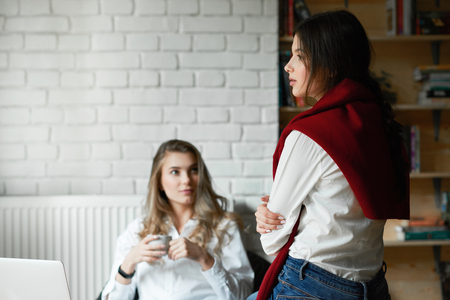Two young female friends wearing casual classic white shirts communicating,working with laptop, sitting in spacy room with white walls. Drinking coffee, learning. Businesswomen, freelancers.の写真素材