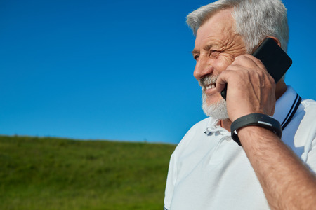 Positive, smiling old man talking on cell phone in green field. Feeling good, satisfied. Wearing white polo shirt with dark blue stripes, sport watch. Standing on saturated sky background.の写真素材