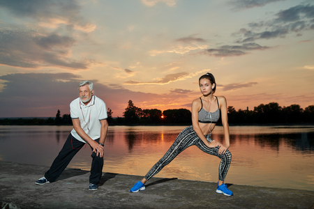 Senior man and young girl having sport training near lake on sunset. Fitness, yoga, crossfit exercises. Fit, strong bodies, healthy lifestyle. Different generations. Outdoors workout on fresh air.の写真素材