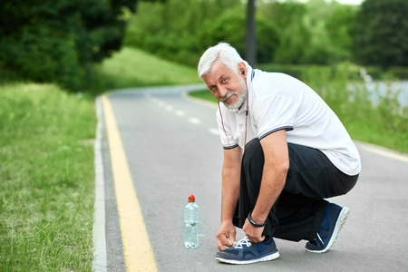 Senior sportsman tie sneakers laces sitting on race track, looking at camera. Keeping body fit, muscular, strong. Maintaining healthy lifestyle. Wearing white t-shirt, black trosers, sport handwatch.の写真素材