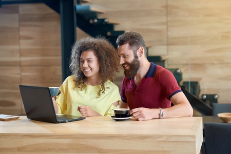 Laughing couple using laptop drinking coffee sitting in cafe. Students posing on wooden table standing in modern restaurant with wooden loft interior. Black metallic stairs on background.の写真素材