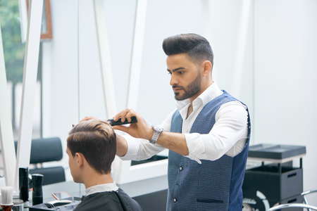 Barber doing new haircut for young client using black plastic comb. Wearing white casual shirt, grey waistcoat, watch. Looking concentrated, loving his job. Model covered with special black cape.の写真素材