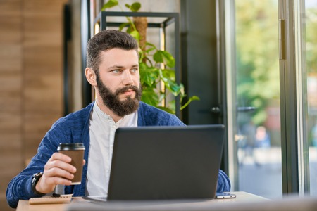 Attractive man with beard and blue eyes working with laptop near big panoramic windows in restaurant with stylish interior. Having dark brown hair bright blue cardigan and white shirt. Drinking coffeeの写真素材