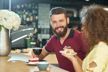 Front view of smiling boy with a beard drinking coffee with curly girl in cafe with modern stylish loft interior. Keeping cup, eating cake, looking at camera. Happy, positive, laughing.の写真素材