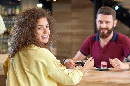 Back view of curly girl sitting in cafe with boyfriend, eating cake, smiling, looking at camera. Feeling happy, satisfied, drinking coffee. Model wearing yellow blouse, man wearing beard, polo shirt.の写真素材