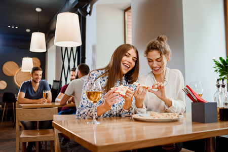 Gorgeous young women sitting in restaurant and having fun. Two best friends eating appetizing pizza and drinking delicious white wine. Girls smiling, looking at camera and posing.の写真素材