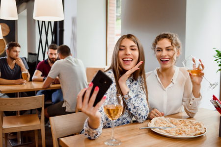 Two best friends making selfie, looking at phone and smiling. Beautiful young women having fun together. Positive girls drinking white wine and having delicious pizza on table.の写真素材