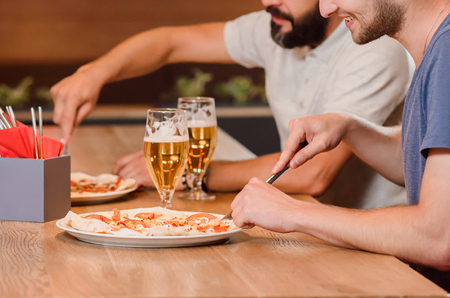 Close up of men cutting appetizing pizza with help of knife and fork. Two male friends also have tasty beer on wooden table. Concept of  pizzeria gastronomy, and cuisine.の写真素材