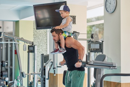 Fit sportsman training on gyms simulator keeping little son on neck. Exercising for healthy lifestyle, having strong hands, muscular body. Dad spending happy family time together with cute child.の写真素材