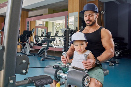 Strong dad and little son doing arms exercises sitting on metallic simulator. Bodybuilder having muscular hands, fit body teaching child maintaining healthy lifestyle, spending family time together.の写真素材