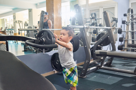 Liitle boy putting heavy dumbbells on metallic simulator standing in gym. Wearing wide colorful shirts, grey tshirt. Father standing on background, keeping healthy lifestyle, having strong body.の写真素材