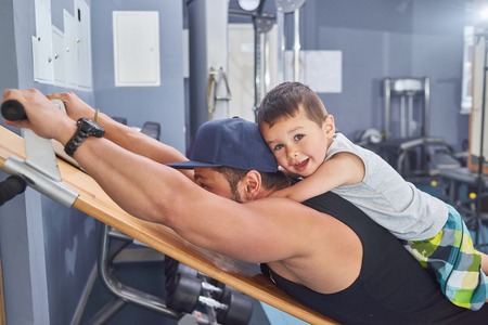 Young father with strong muscles training in gym keeping little son on his back. Wearing sport tshirt, cap. Child smiling, laughing, feeling happy, looking at camera. Family time together.の写真素材