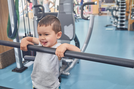 Little cute boy grimacing, holding on metallic crossbar in gym. Child laughing, smiling, looking cute, happy, satisfied. Heavy simulators standing behind. Keeping healthy lifestyle.の写真素材