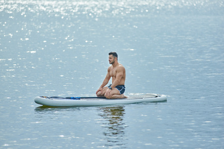 Sporty man sitting on sup board during trainining on lake, looking forward. Having fit, healthy,...