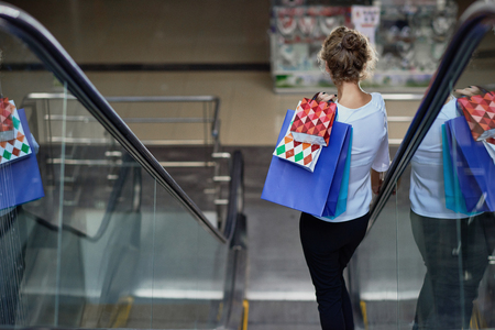 View from back of female customer with shopping bags driving down on escalator in mall. Curly lady enjoying good shopping and new purchases. Concept of buying and pleasure.の写真素材