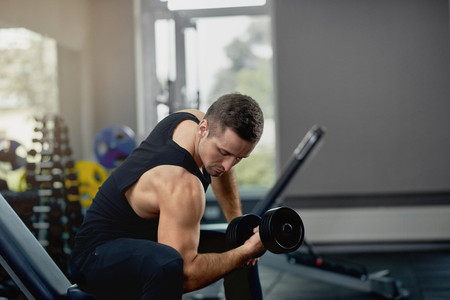 Handsome sporty man flexing muscles with barbell in dark gym.の写真素材
