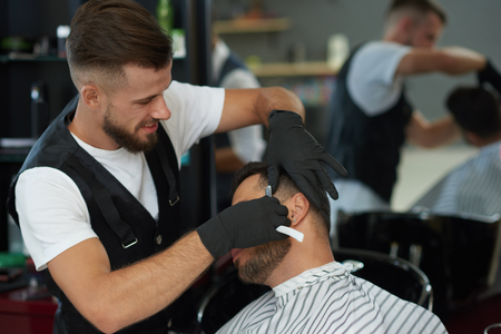 Professional barber holding razor in hands and shaving face of man. Male hairdresser wearing black...