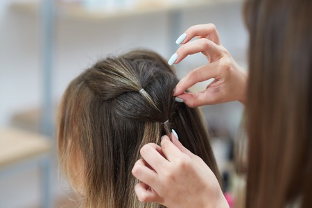 Selective focus of hands of professional hairdresser braiding spikelet to female client in beauty salon. Qualified woman carefully correcting hairstyle. Concept of look preparation.の写真素材