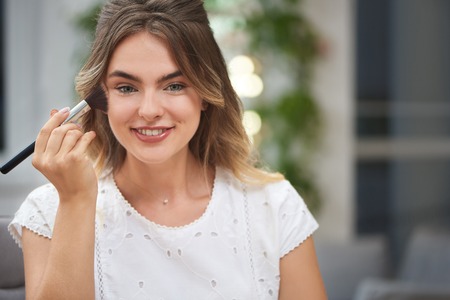 Portrait of charming young girl keeping brush and fixing makeup with powder in salon. Beautiful female with trendy hairstyle looking at camera, smiling and posing. Concept of beauty and visage.の写真素材