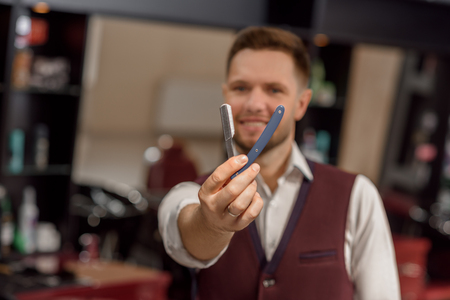 Male hairdresser looking at camera and holding instrument for shaving hair. Razor in hands of barber posing on background. Concept of barbershop and styling of hair.の写真素材