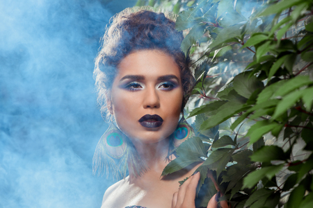 Portrait of sexy sensual girl with dark purple lips, bright eyes and earrings made of peacock feathers. Woman with bare shoulder, provocative posing outside against green bushes in light blue smoke.の写真素材