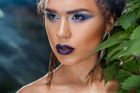 Close up of seductive young girl with bright colored makeup and peacock feathers earrings, looking at side. Gorgeous hot woman with bare shoulders covered with sparkles after salon posing outside.の写真素材