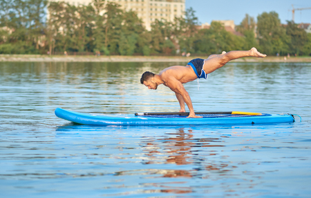 Muscular man practicing yoga, doing balance body weight, swimming on paddle board in middle of city lake, during sunny summer day. Sportsman with perfect figure, wearing blue shorts, training outside.の写真素材