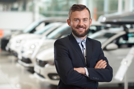 Front view of handsome man standing in modern car center and posing. Bearded manager wearing in dark blue jacket smiling and looking at camera. Row of automobiles on background.の写真素材